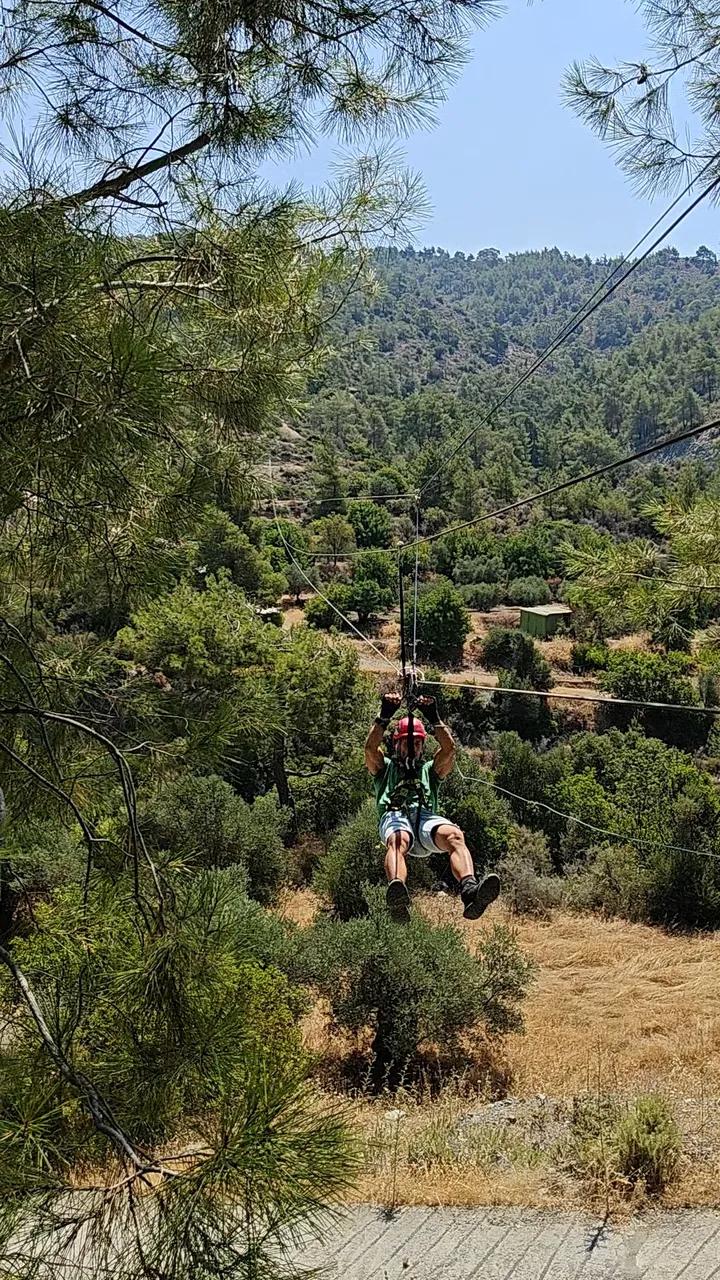 Person in a red helmet and harness zip-lining toward the camera, gripping the trolley, forest valley in the background.