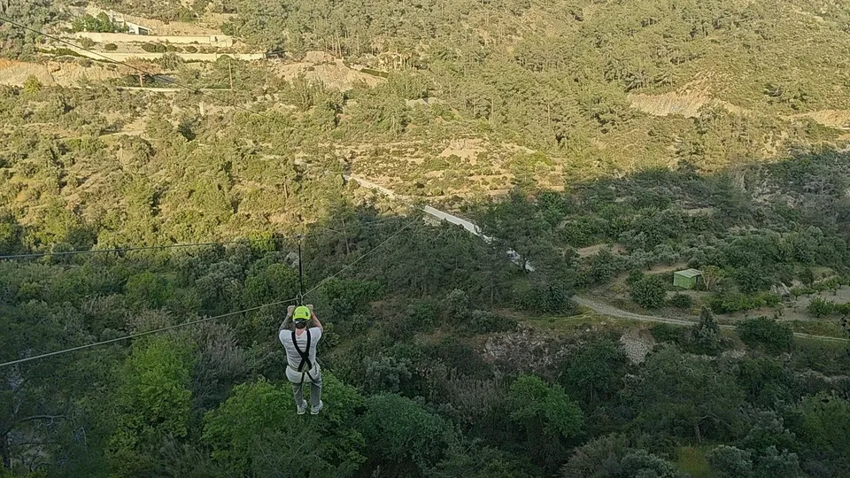 Person in helmet and harness zip-lining over a forested valley, seen from behind, hills and greenery below.