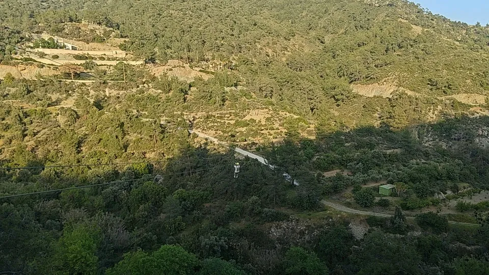 Wide view of a pine-forested valley with a person suspended on a zipline cable, a road and green shed below.